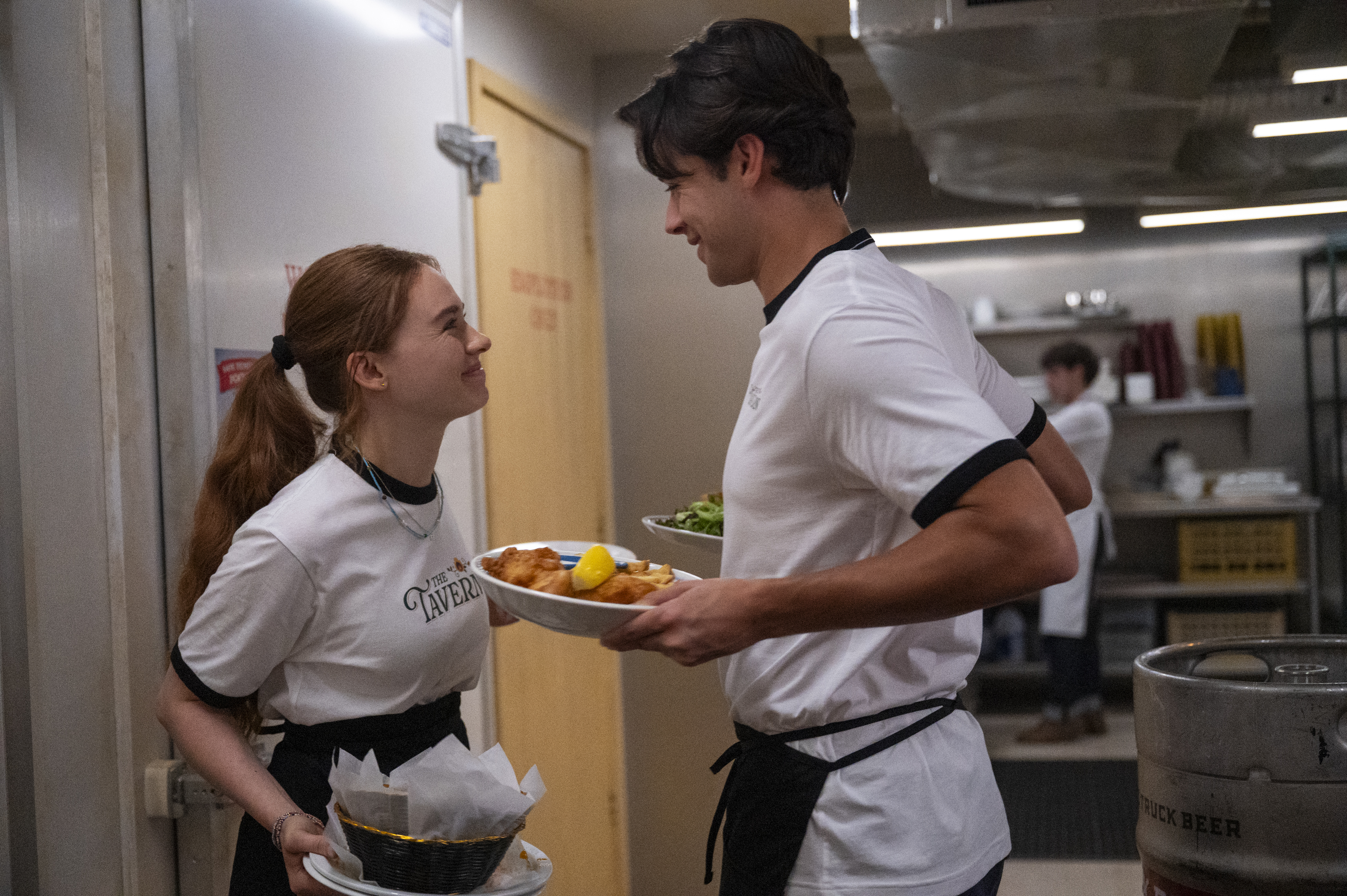 a young woman and young man wearing matching uniforms and holding food while working in a restaurant kitchen in a still from every year after