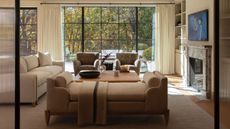 A bright, airy living room viewed through fluted glass doors, featuring a large black-framed window that overlooks lush autumn trees. The space is anchored by a neutral sofa and two textured charcoal armchairs centered around a wooden coffee table, with a marble-surrounded fireplace and built-in shelving on the right