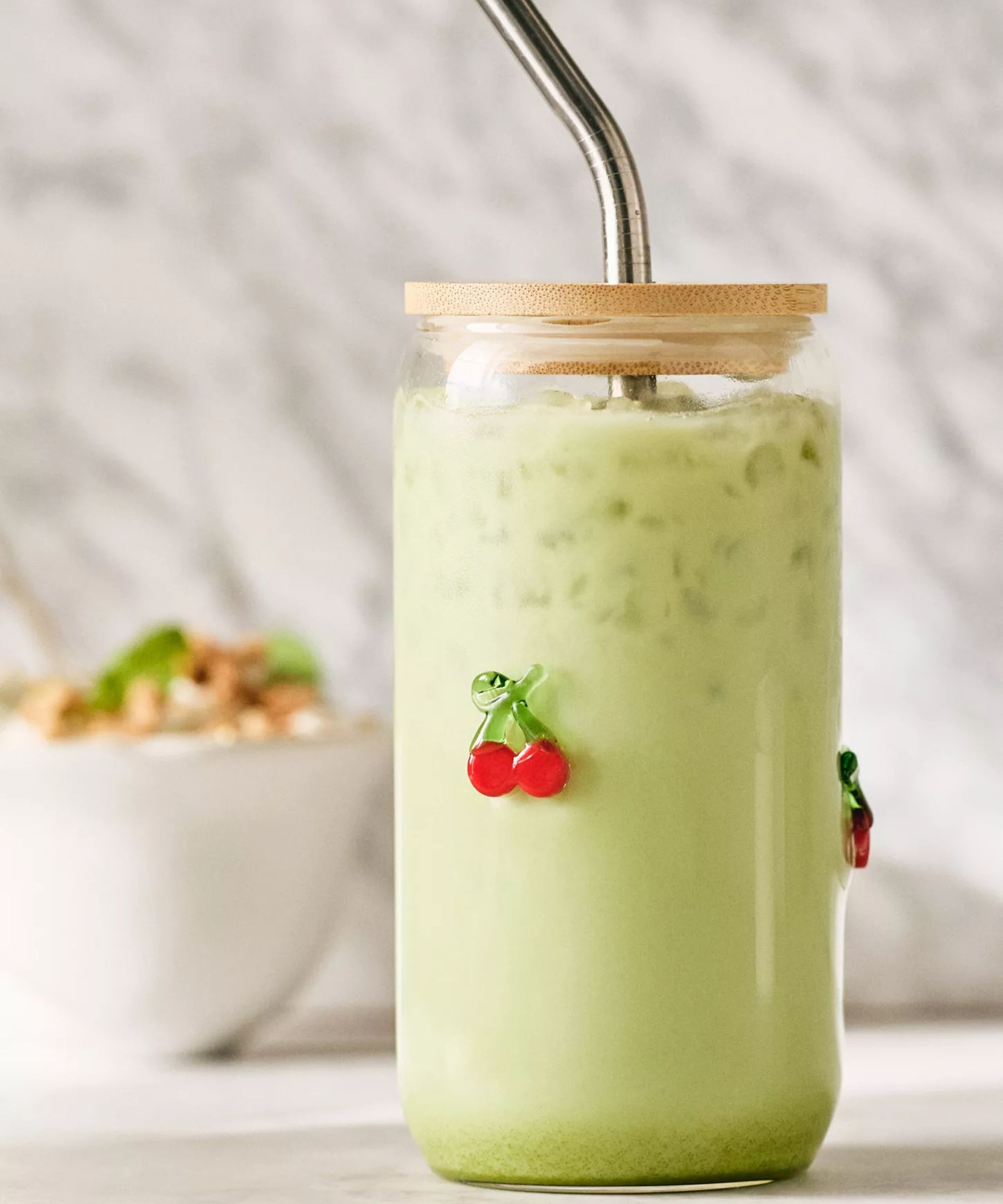 Glass travel cup with wooden lid and metal straw and glass cherry embellishment. Inside is a pale green drink with crushed ice floating at the top