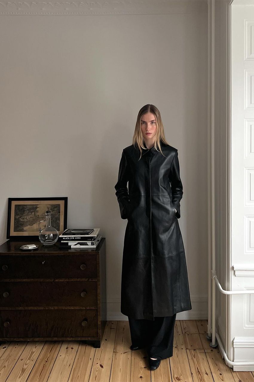 A woman standing inside an apartment by a wood dresser that's decorated with art and books. She's wearing a longline leather duster jacket with black wide-leg pants and black ballet flats.