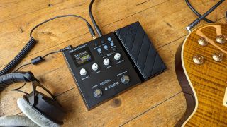 An NUX MG-300 multi-effects pedal on a wooden floor with a pair of headphones and electric guitar