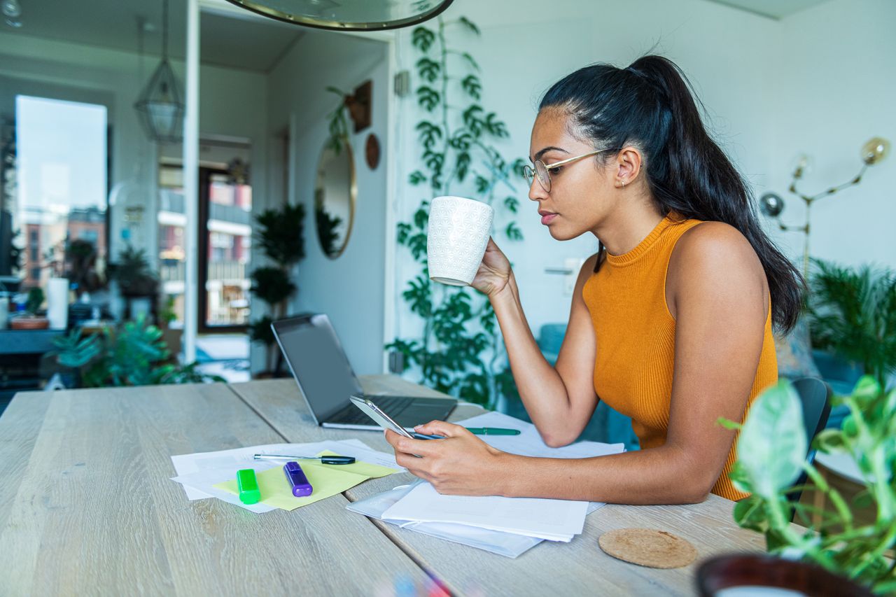 A young woman drinks coffee while looking at personal finance information on her phone.