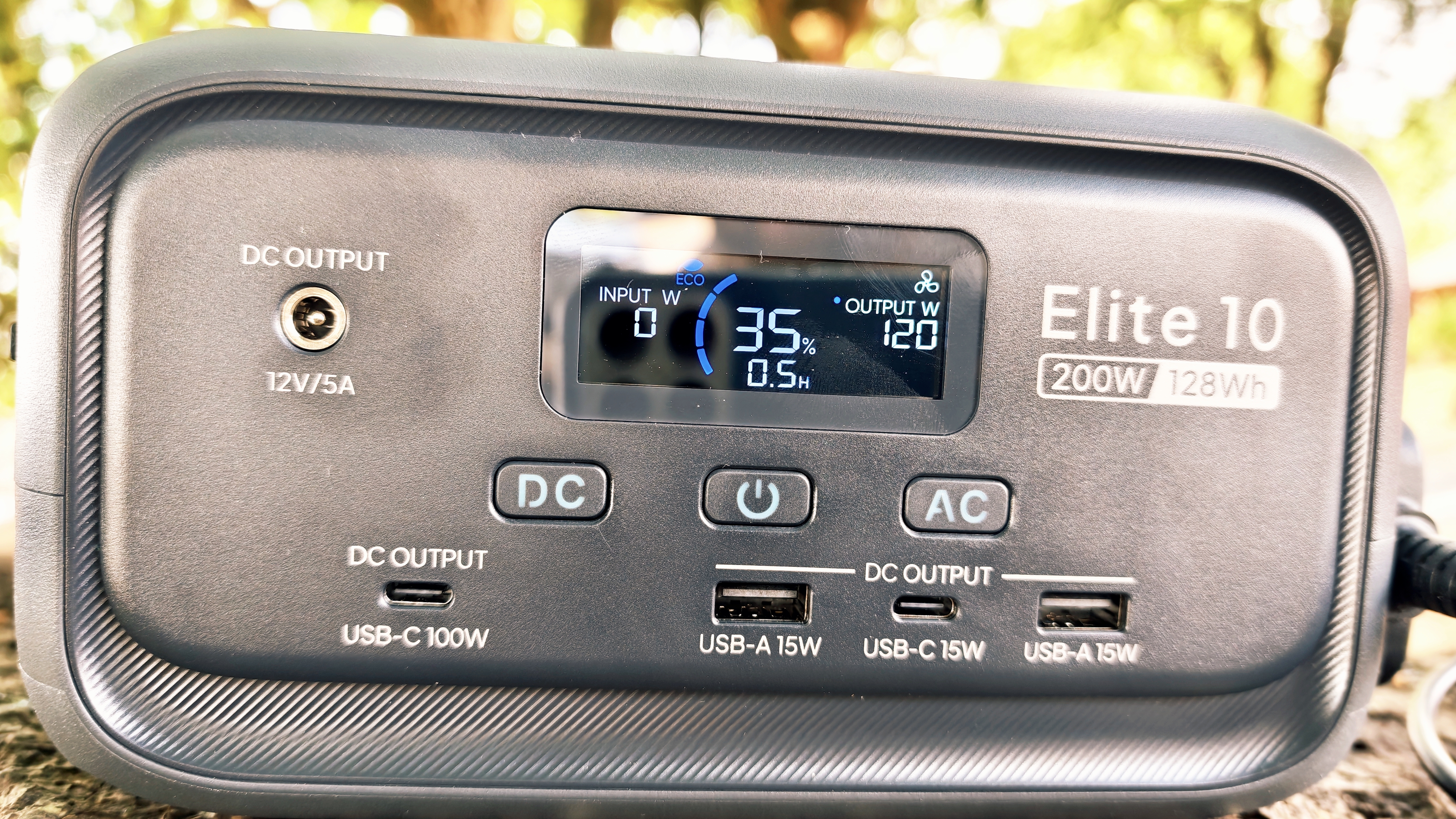 A grey Bluetti Elite 10 power station sitting on a log in a forest grove