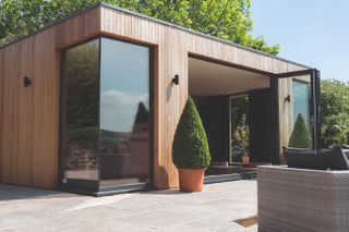 a modern wood panelled garden room with floor-to-ceiling windows and sliding doors, garden furniture outside, and a large plant by the door