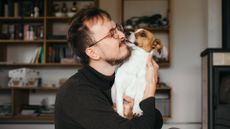 A man wearing glasses and a black roll-neck jumper is shown being licked by his dog on the face. 