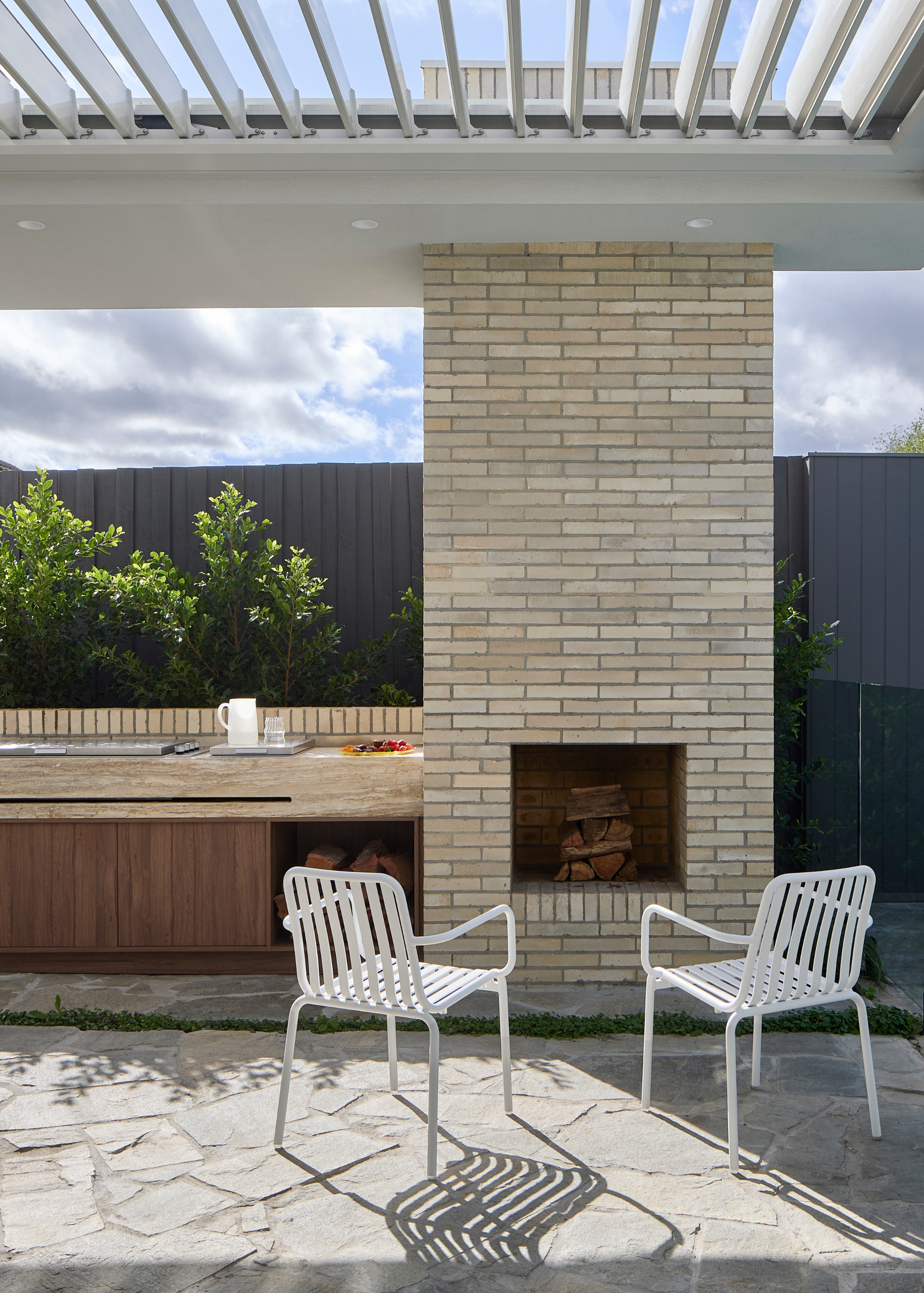 An outdoor space with a pair of white metal chairs in front of a fireplace, near an outdoor kitchen
