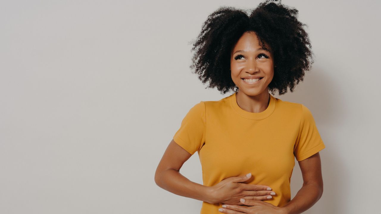 A happy woman in a yellow t-shirt rubs her belly in front of a plain background