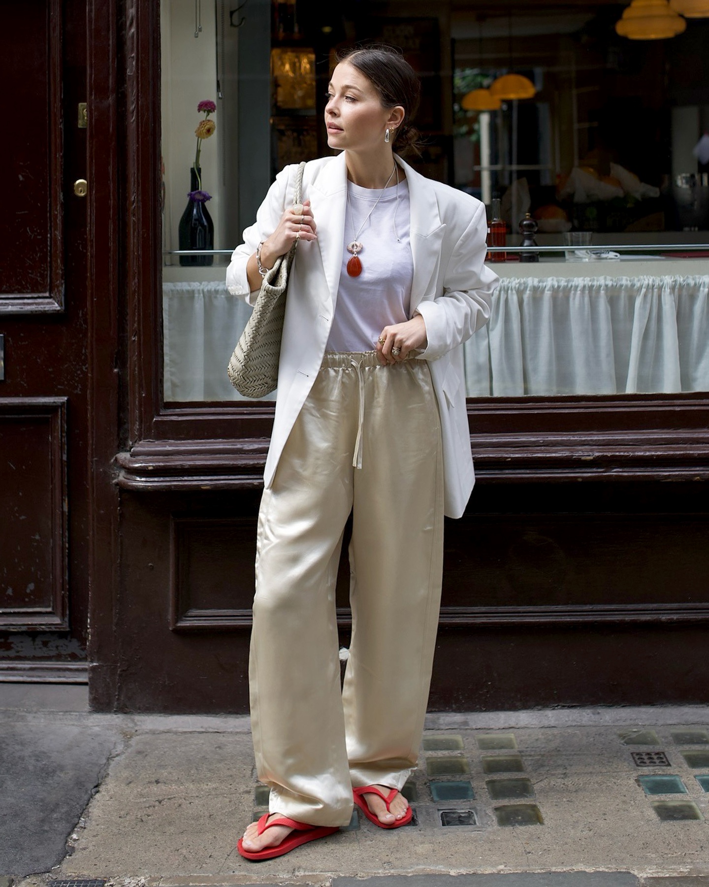 British style influencer Alice Satterthwaite poses in London wearing a neutral blazer, white T-shirt, red stone pendant necklace, ivory woven tote bag, beige satin drawstring pants, and red flip-flops
