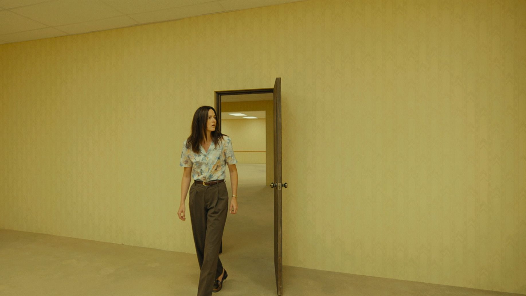 A pale yellow film set with textural wallpaper, minimal decor, and people wandering through it.