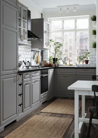 grey kitchen cabinets and drawers. Glass kitchen cabinets have been places on the upper levels of the kitchen. There is also a stove and cooking essentials hanging. There are also a few plants by the window.