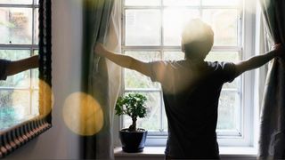 Man standing at the window opening curtain to let morning light in.