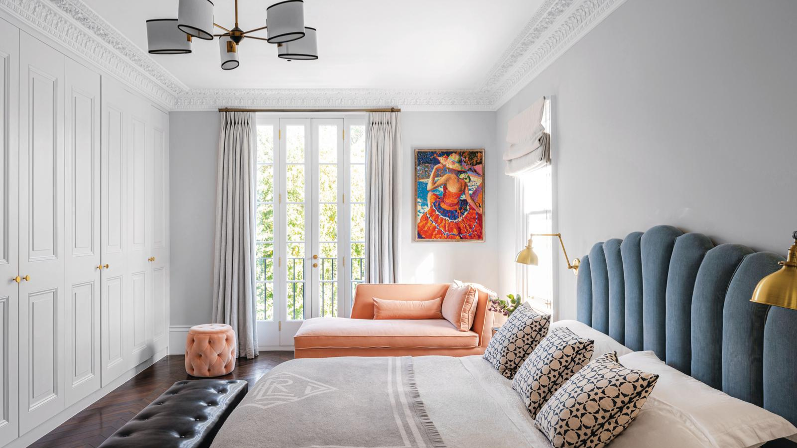 a bedroom with a white ceiling and walls, a large bed with a blue headboard, a salmon pink chaise lounge, and colorful artwork on the wall