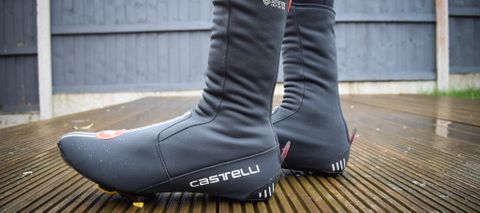 A close up of a black pair of Castelli Estremo overshoes worn by a cyclist who is stood on a wooden deck
