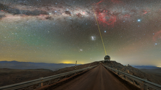 A telescope sits underneath a speckled, colorful night sky as a beam of laser light shoots up as a laser star guide.