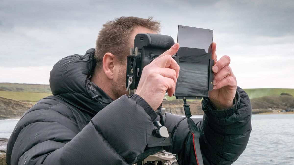 Photographer adjusting a neutral density filter on a tripod-supported camera in a coastal landscape