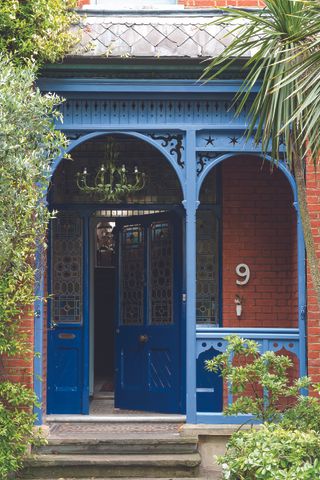 A red brick house surrounded by greenery and an ornate blue porch painted in Farrow and Ball Pitch Blue