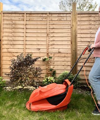 A woman wearing light pink cardigan, light blue denim jeans and black casual sneaker footwear mowing lawn with lawnmower with fence in background