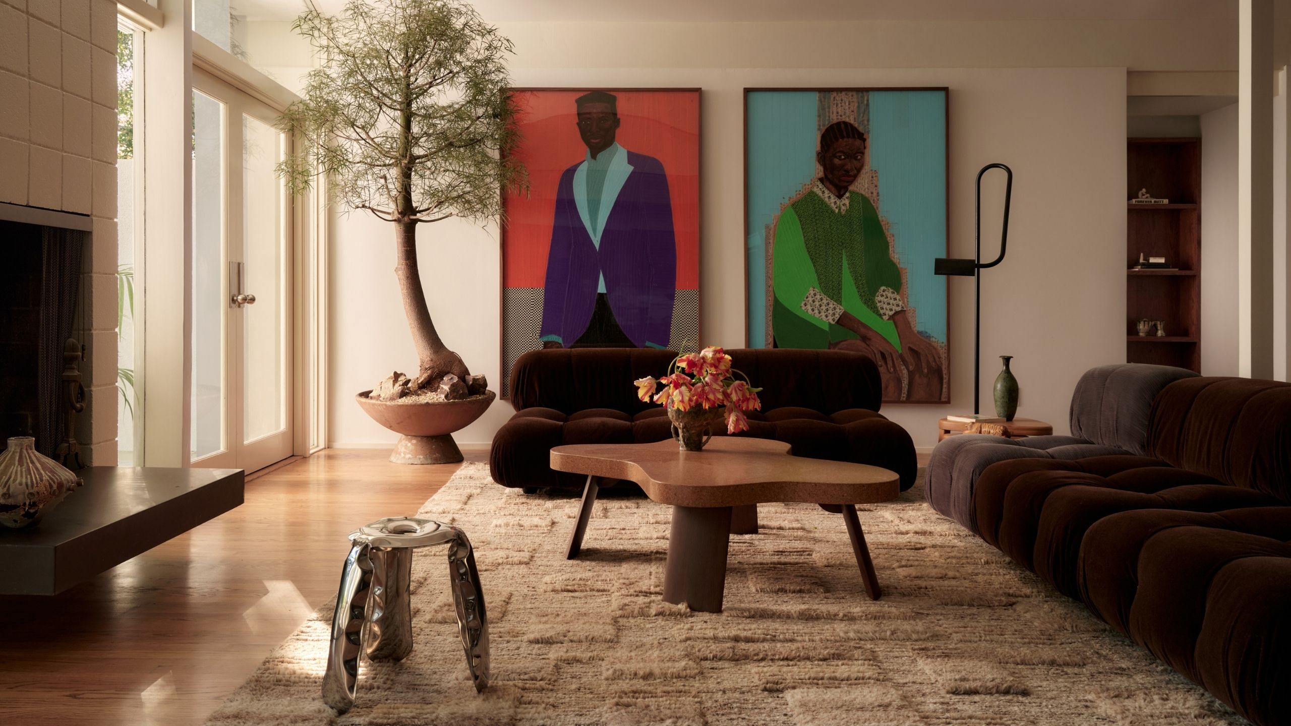 A beige and brown-tinted living room with modular, softly shaped sofas, large figurative canvases, a tall potted plant, and a textured rug alongside wooden and chrome furniture.