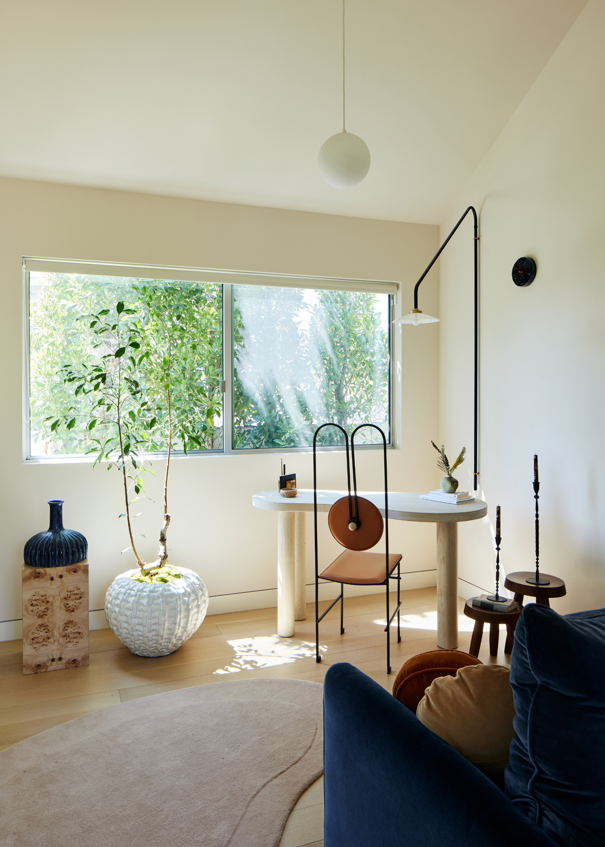 A white study space with a minimalist desk by the windows, pendant lighting, a statement chair, a potted plant and a vase on a burl wood plinth by a rug near a pair of wood stools with candlesticks and a blue sofa seat