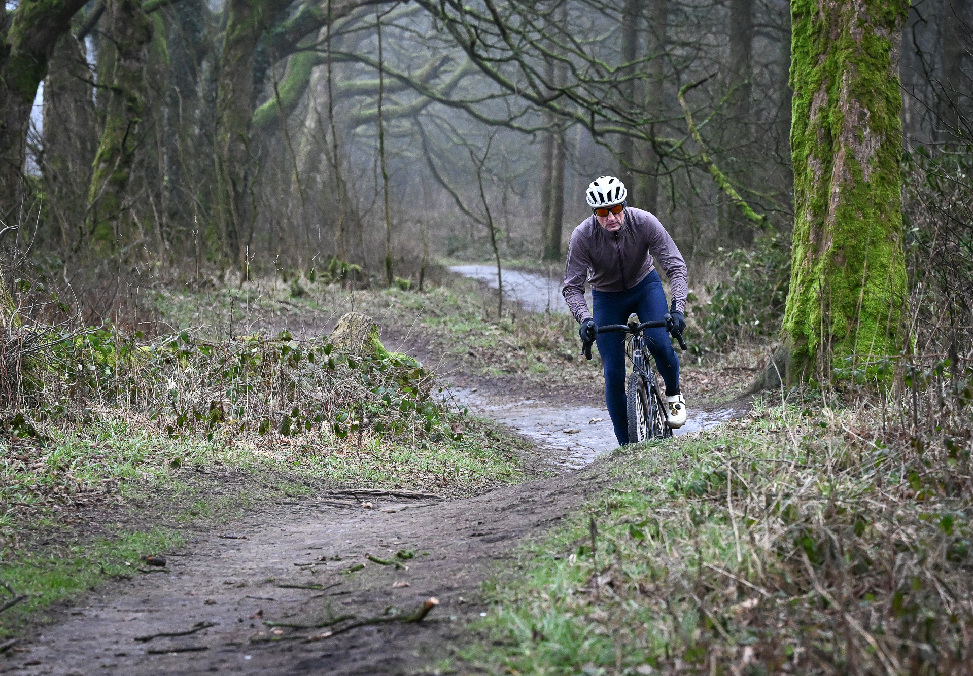 Man riding a gravel bike on a dirt track wearing blue tights, purple jacket and beige shoes