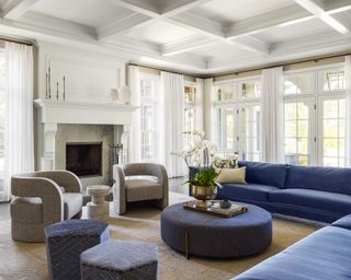 Large living room with blue corner sofa, two gray armchairs, round blue ottoman, fireplace, white painted walls, beige rug
