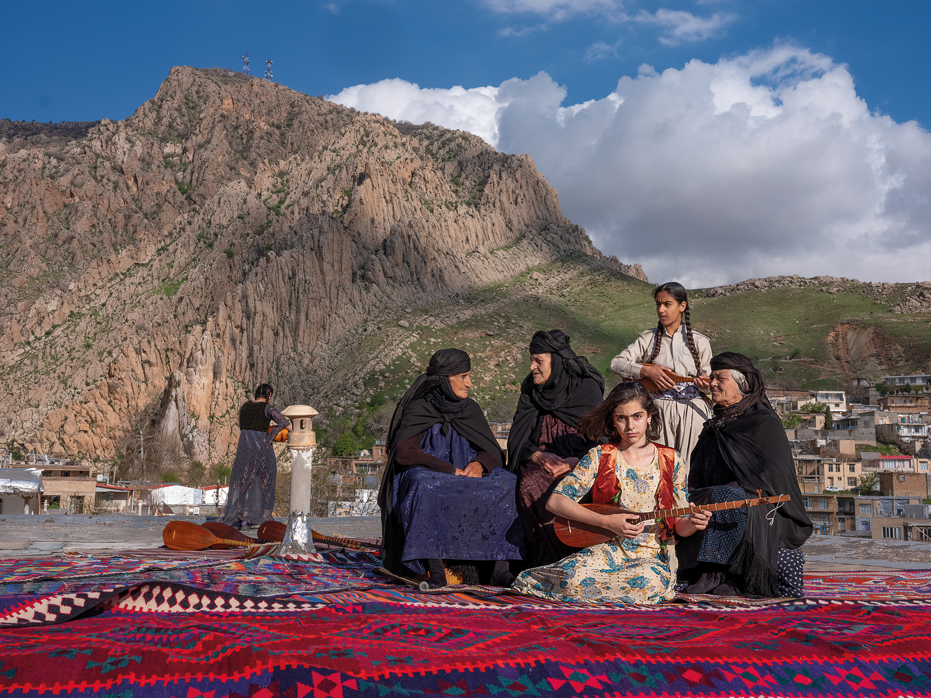 A group of women in traditional clothing plays musical instruments on a colorful rug, with a mountainous landscape and village in the background
