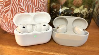 The Apple AirPods Pro 3 and Sony WF-1000XM5 side by side on a wooden surface in their charging cases.