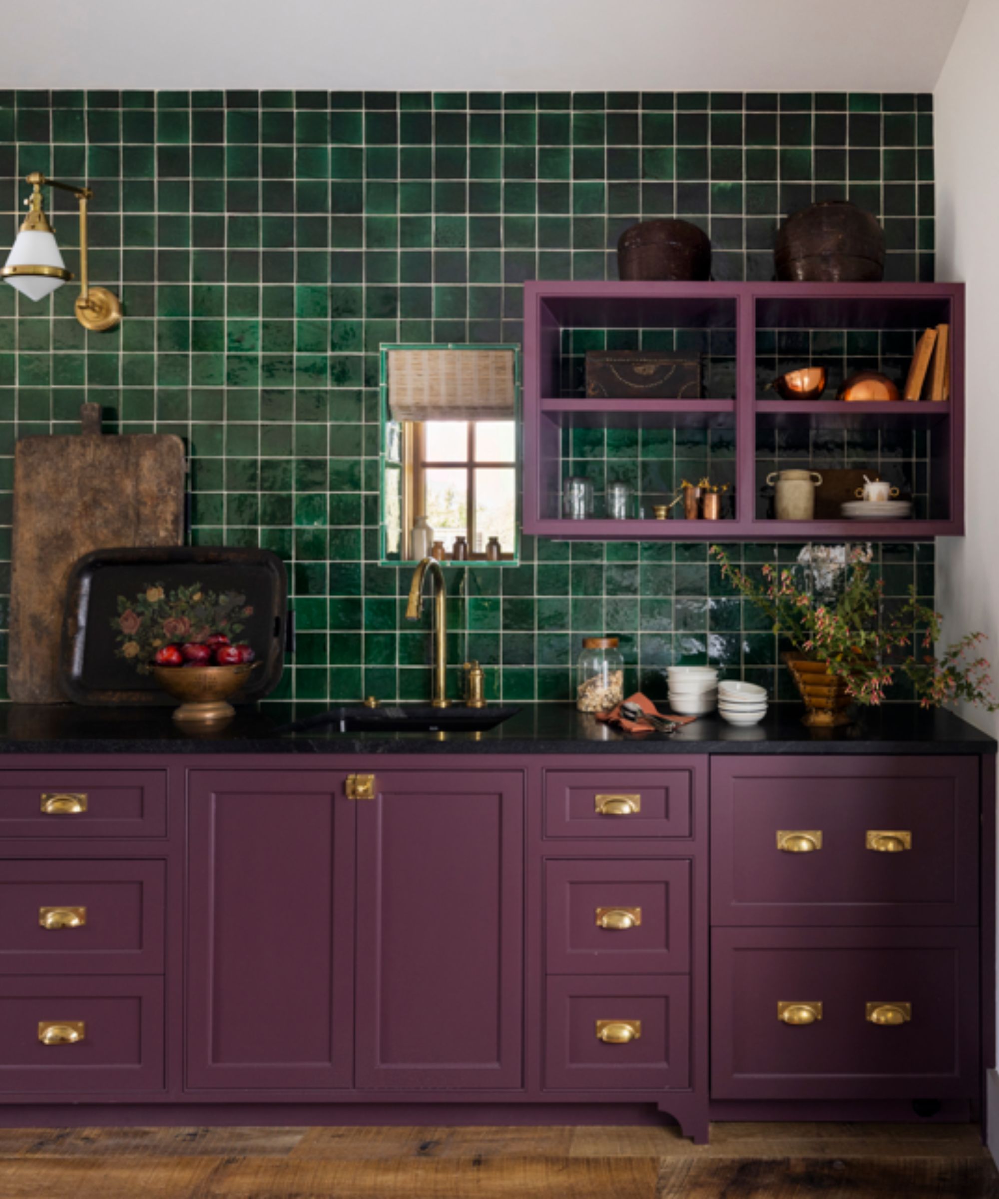 Kitchen with aubergine cabinetry and gold handles, green tiled wall, open shelving with ceramics and glassware, black marble countertops, and a large wooden chopping board.