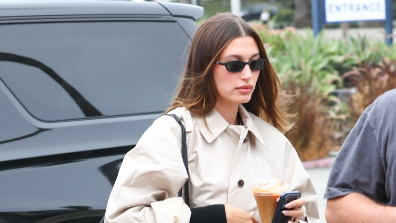 Hailey Bieber wearing a trench coat, black flare pants, and plain black leather handbag at the DMV