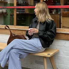 British influencer Chloe Butler sitting on a bench outside a London café holding a coffee wearing an oversize black leather bomber jacket, a neutral sweater, striped blue pajama pants, and a tan leather bag