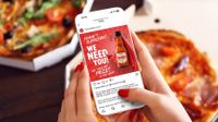 A person holds a smartphone displaying a social media contest page for Frank's RedHot sauce, set against a background of fresh pizzas.