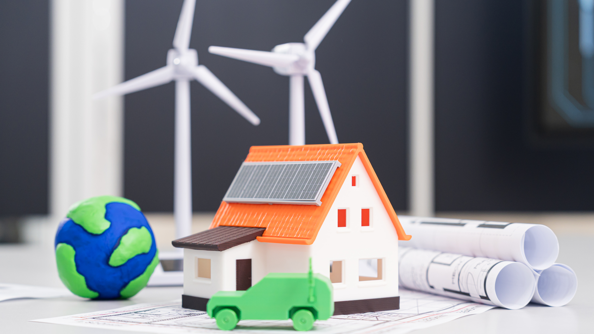 model of house with solar panels, model of wind turbines, green toy car, rolled up architectural drawings and model of earth all on desk