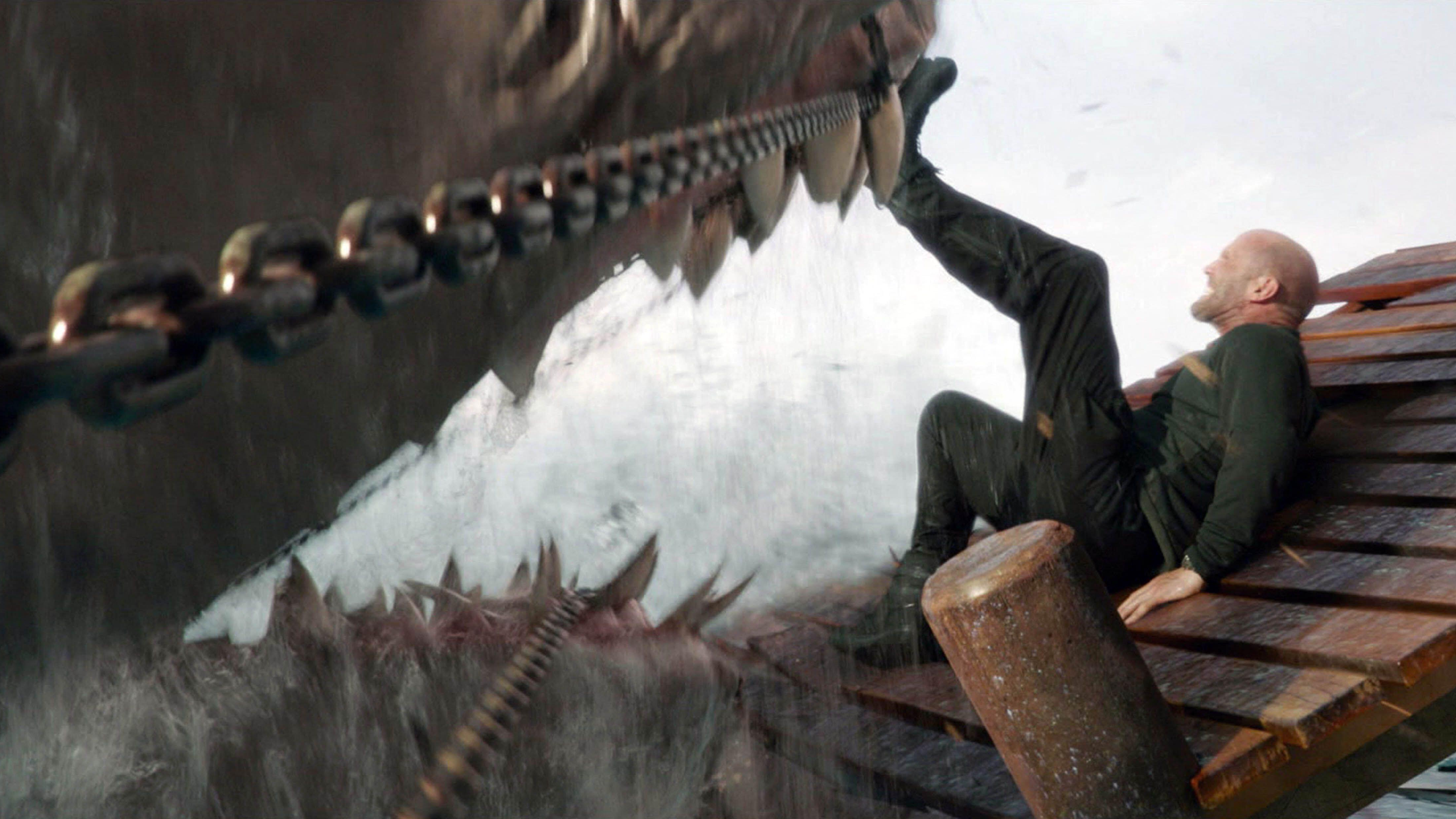 Jonas Taylor (Jason Statham) lies on a pier holding a hungry chained megalodon shark back with his feet in a scene from "Meg 2: The Trench"
