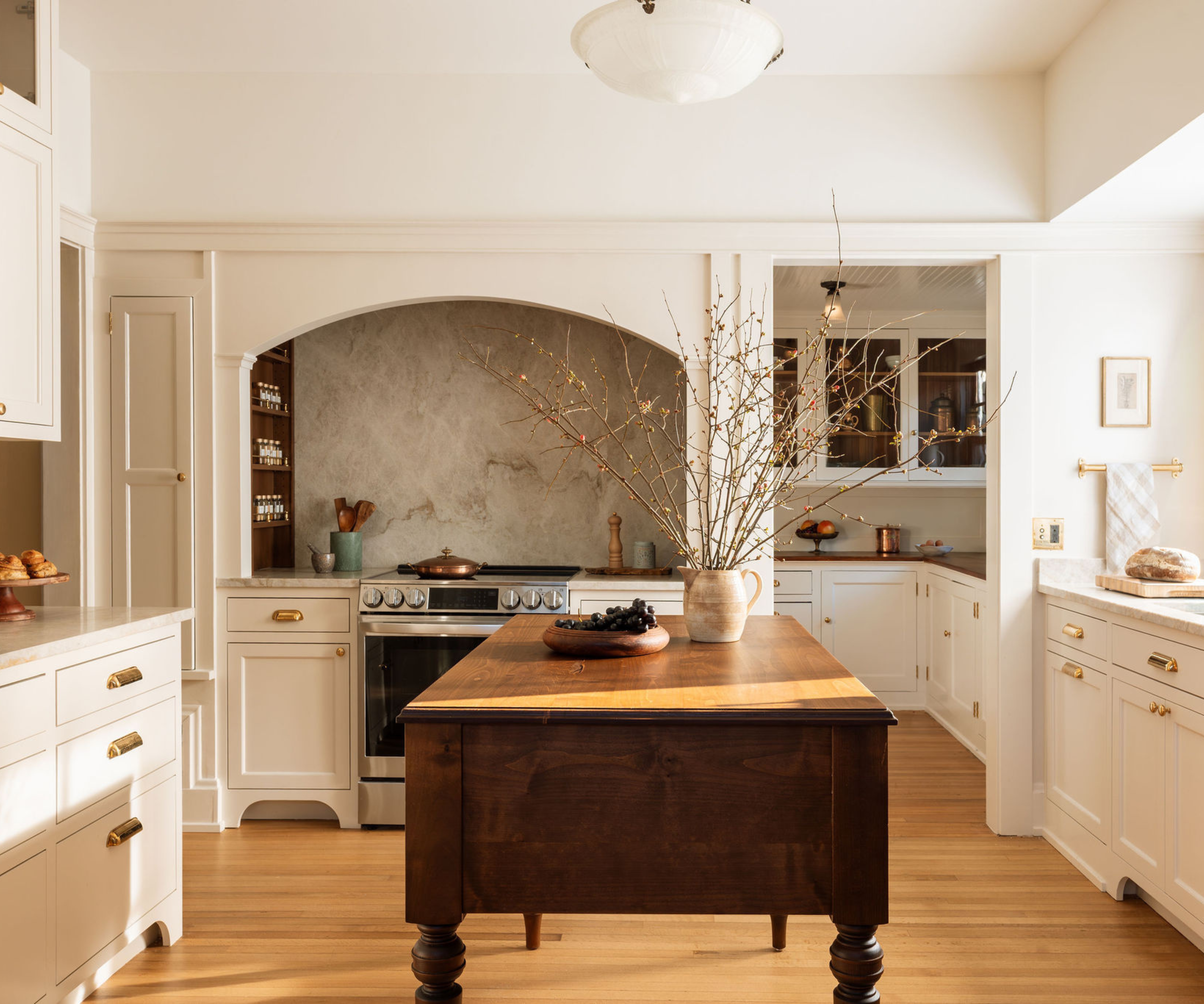 A warm, traditional kitchen featuring a large arched alcove over the stove with a textured stone backsplash, off-white cabinetry with brass bin pulls, and a dark wood antique-style island in the center