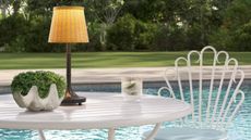 Portable wicker outdoor lamp aglow atop of a white patio set overlooking a pool and grassy lawn