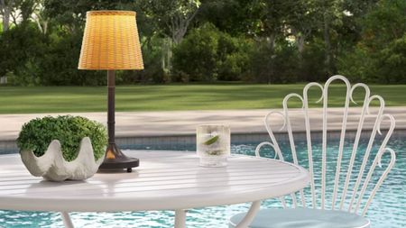 Portable wicker outdoor lamp aglow atop of a white patio set overlooking a pool and grassy lawn