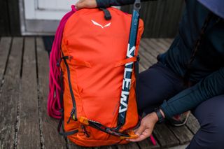 A close-up of a bright orange Salewa Ortles Wall 38L Backpack, showing an ice axe being fitted into place.