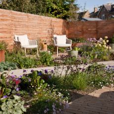 a garden with two armchairs in the sunshine beside paths with plenty of flowers and plants