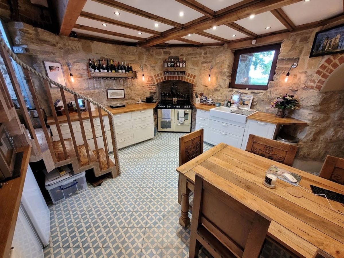 The kitchen has tiled floors with wooden beams along the ceiling and a staircase leading out with windows showing out onto the garden with a oak wooden table and chairs which have a TV next to it on the adjacent wall. The cupboards are white with wooden worktops