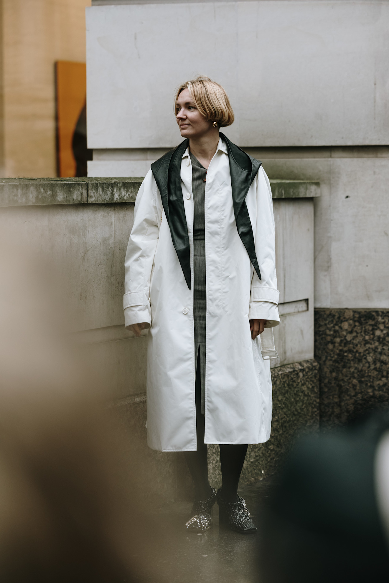 a woman at london fashion week wearing a black leather triangle scarf, white coat, grey dress, and crystal booties