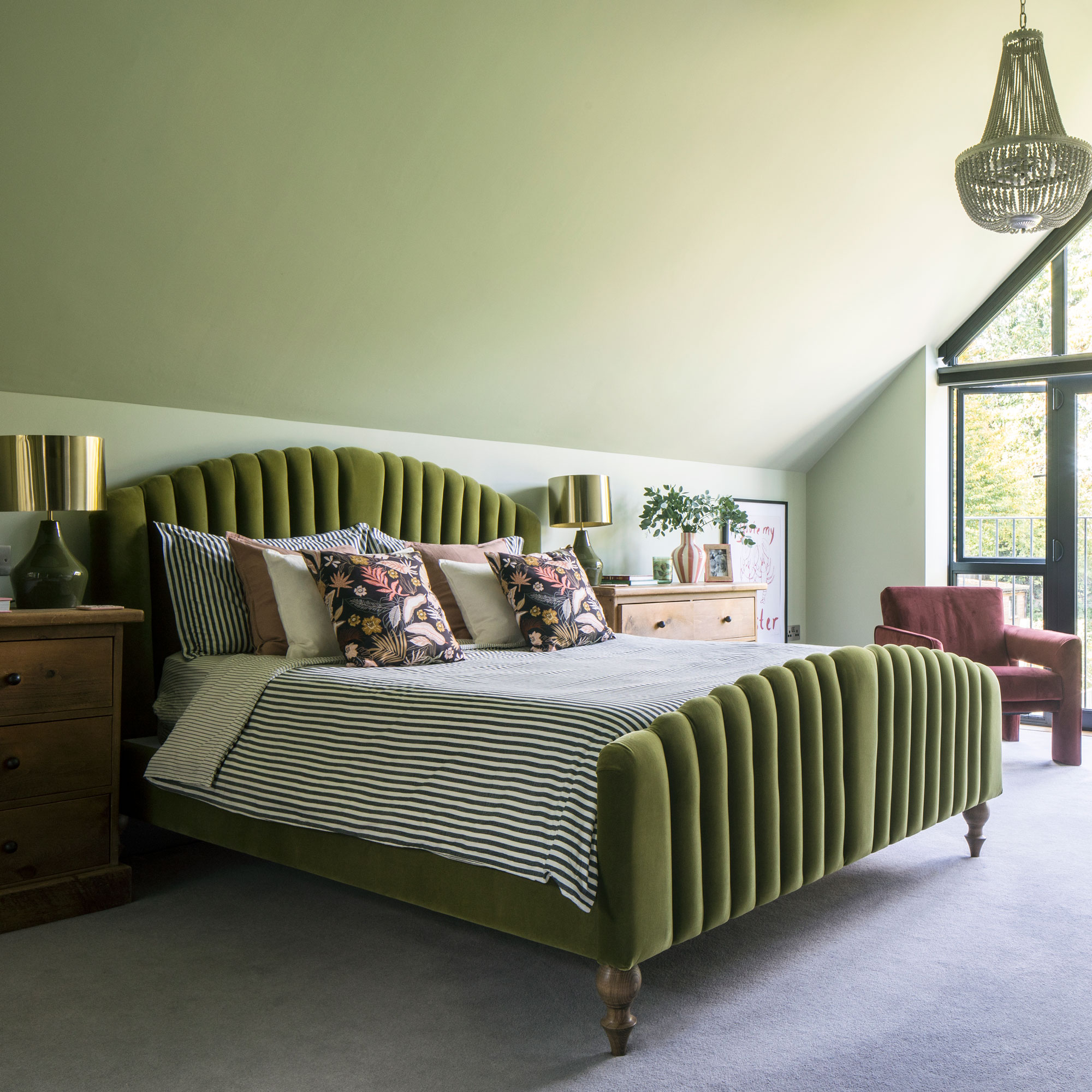 Green velvet double bed in a green colour drenched room with a chandelier, wooden side boards and pink armchairs