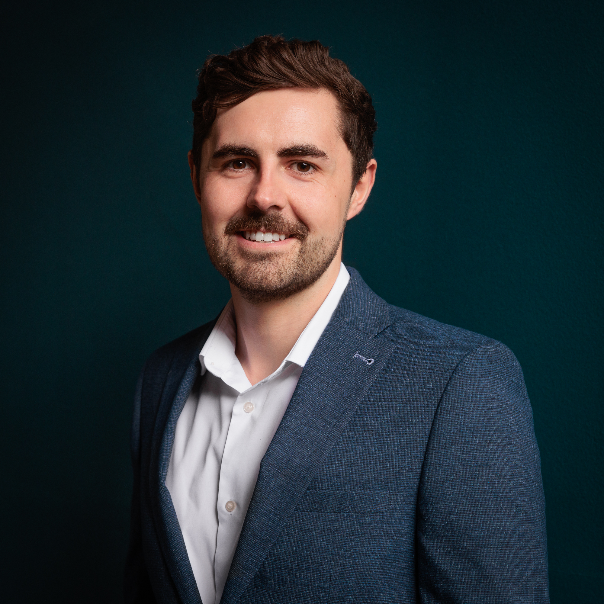 male with short brown hair, facial hair wearing blue suit jacket and white shirt