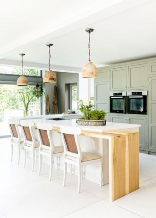 kitchen island with white worksurface and white painted upholstered chairs with green cabinets behind and double oven