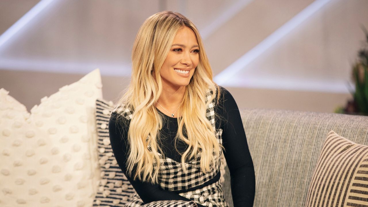 Hilary Duff in a black top and checked dress sitting on a gray couch with pillows on it