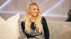Hilary Duff in a black top and checked dress sitting on a gray couch with pillows on it