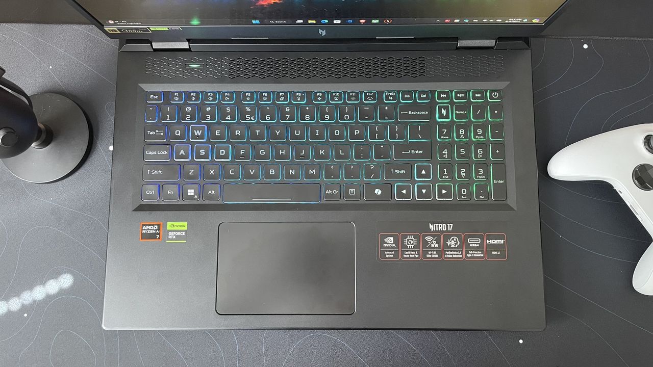 The backlit keyboard deck of the Acer Nitro 17, on a black desk with a white controller to the right.