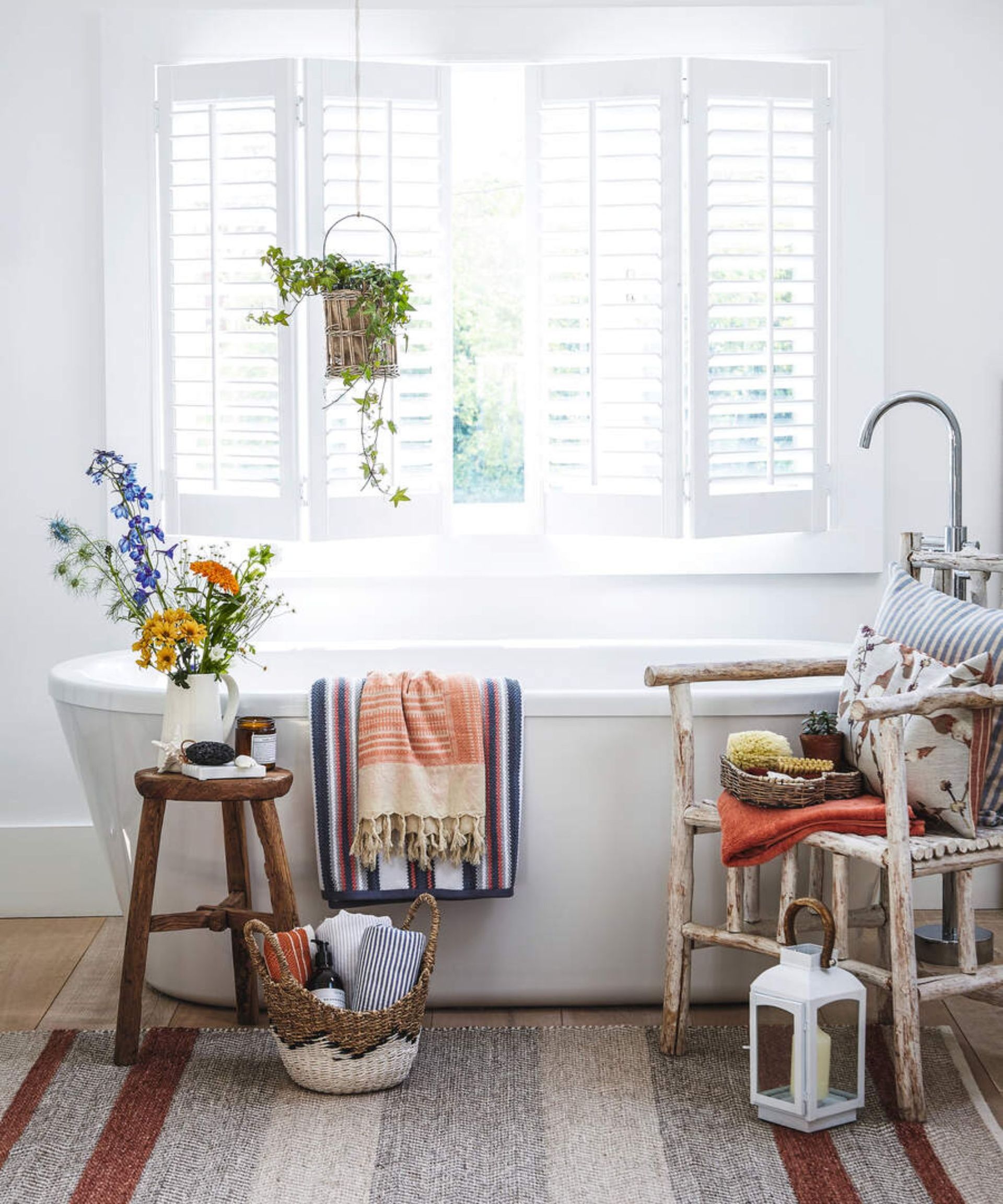 A bright white bathroom with open windows and white shutters, a white standing bath, and a striped rug. There is a wooden chair with cushions, and a wooden stool opposite it, but with white jug filled with colorful flowers. On the floor is a white and brown basket filled with rolled towels, and a white lantern with a candle. Hanging on the bath are two striped towels in orange, red and blue. Above the bath is a green hanging plant in a wicker pot.