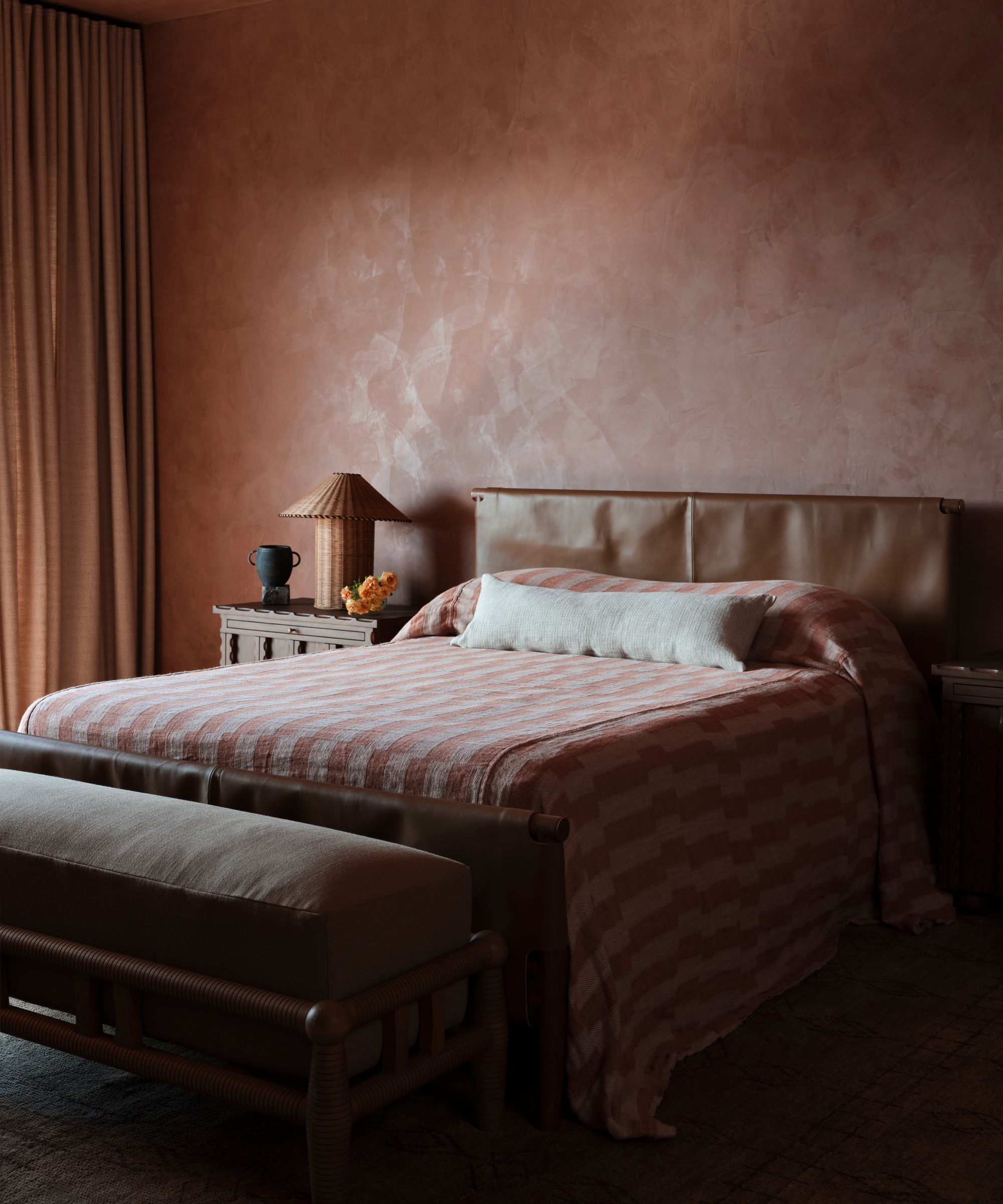 Tonal bedroom with plaster-pink walls and ceiling, striped bedding, leather bed frame, full-length curtains and a woven bench.