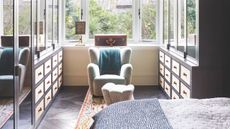 Bedroom with adjoining dressing room with mirrored closets and blue armchair, with large windows and leafy garden seen beyond.