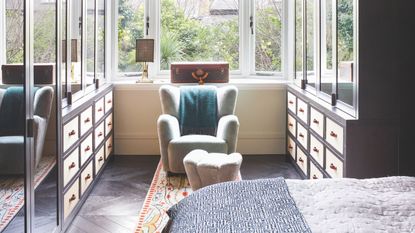 Bedroom with adjoining dressing room with mirrored closets and blue armchair, with large windows and leafy garden seen beyond.